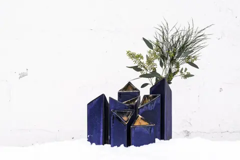 A cluster of tall deep-blue glazed vases of different heights with angled openings, holding greenery, set in snow against a white wall