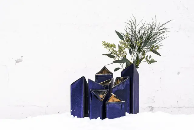 A cluster of tall deep-blue glazed vases of different heights with angled openings, holding greenery, set in snow against a white wall