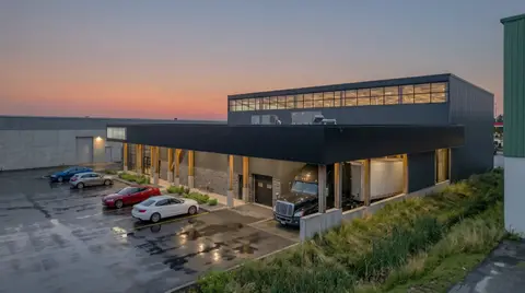 Visualization at dusk of the finished building, with a dark metal-clad warehouse addition, clerestory windows and a timber-post canopy over the entrance and loading dock