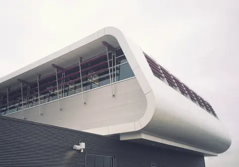 Close view of the building's rounded white cantilevered upper floor, with angled sunshades over its windows, above dark metal cladding