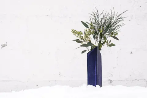 A single tall blue glazed vase with an angled opening, holding eucalyptus and grasses, standing in snow